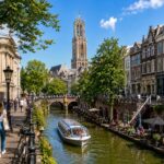 Zonnig uitzicht op de Utrechtse grachten met rondvaartboot, terrassen, historische gebouwen en de Domtoren op de achtergrond